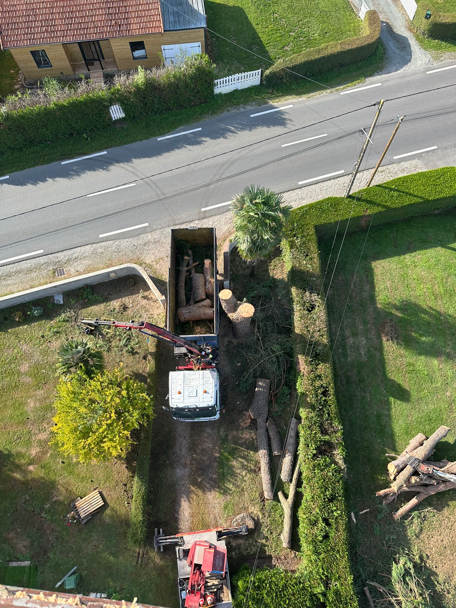 Vue aérienne d'un jardin en bord de route. Des branches et des troncs coupées attendent d'être évacuées par un camion bleu déjà chargé de troncs.