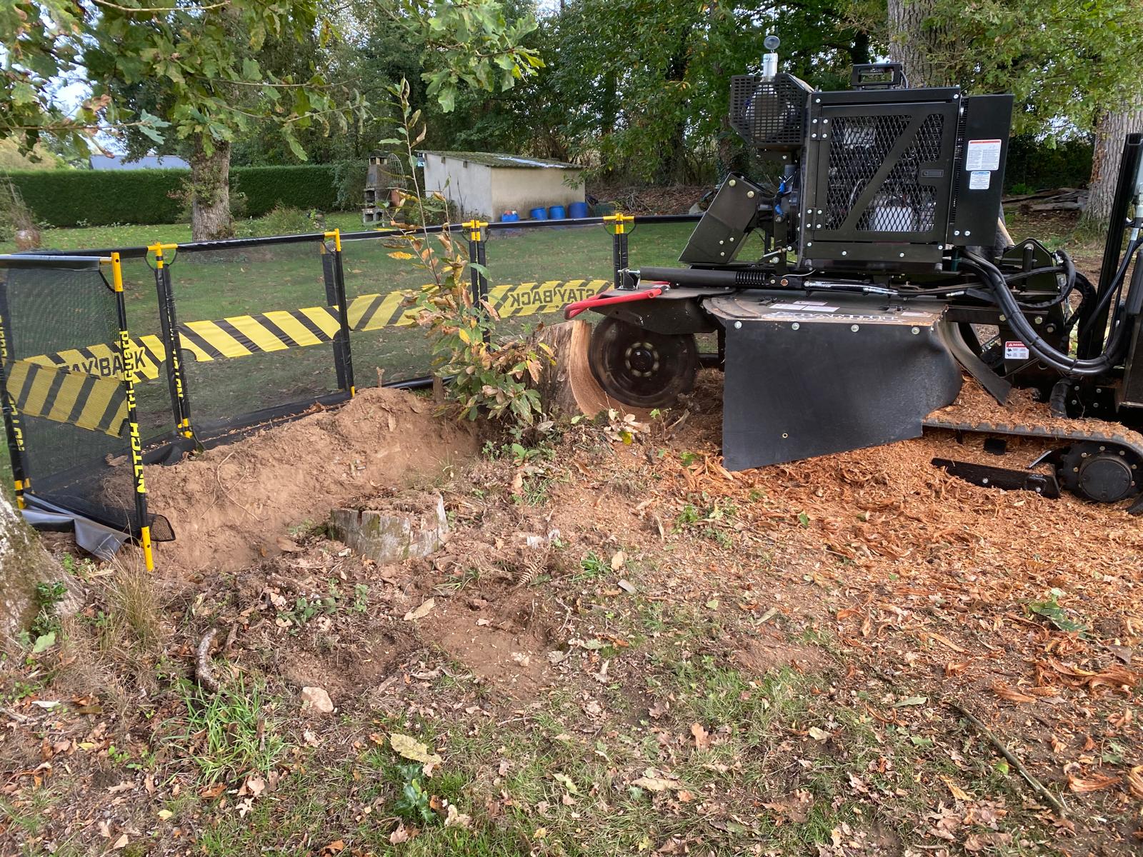 Une rogneuse entourée d'un ruban de sécurité jaune et noir ronge une souche. Des copeaux de bois jonchent le sol autour de la machine.