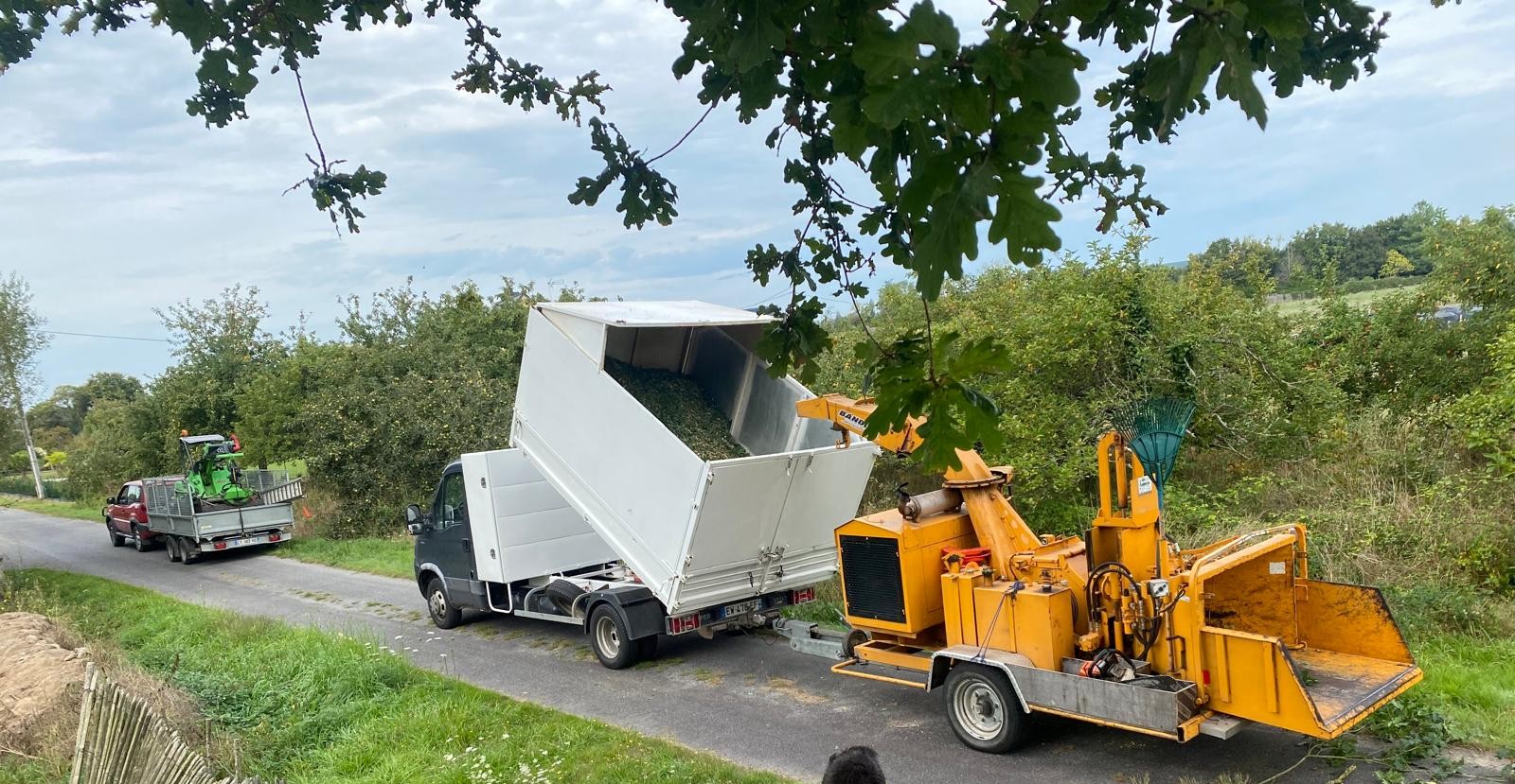 Un broyeur jaune monté sur remorque avale des branches d'arbre en bord de route. Une benne blanche est attelée juste devant pour récupérer les copeaux.